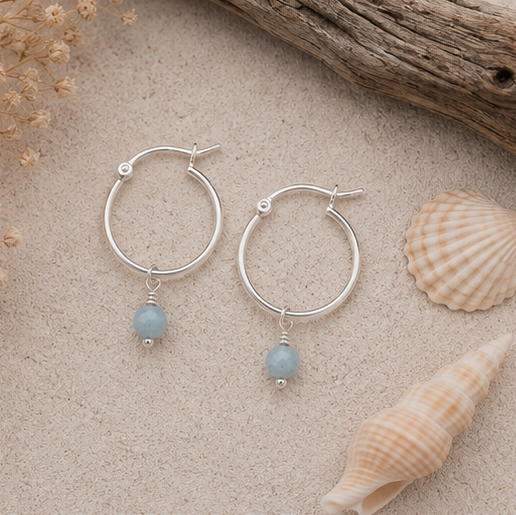 A pair of silver hoop earrings with a aquamarine gemstone bead dangle, displayed on a sandy surface with seashells and driftwood.