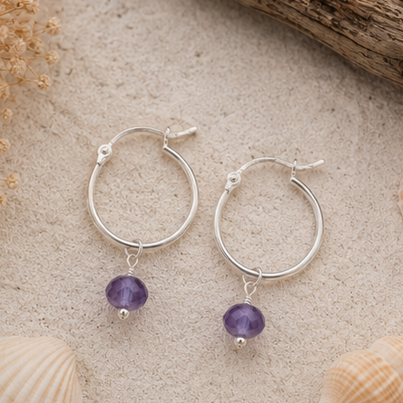 A pair of sterling silver hoop earrings with dangling purple amethyst beads, displayed on a sandy surface with seashells and driftwood.