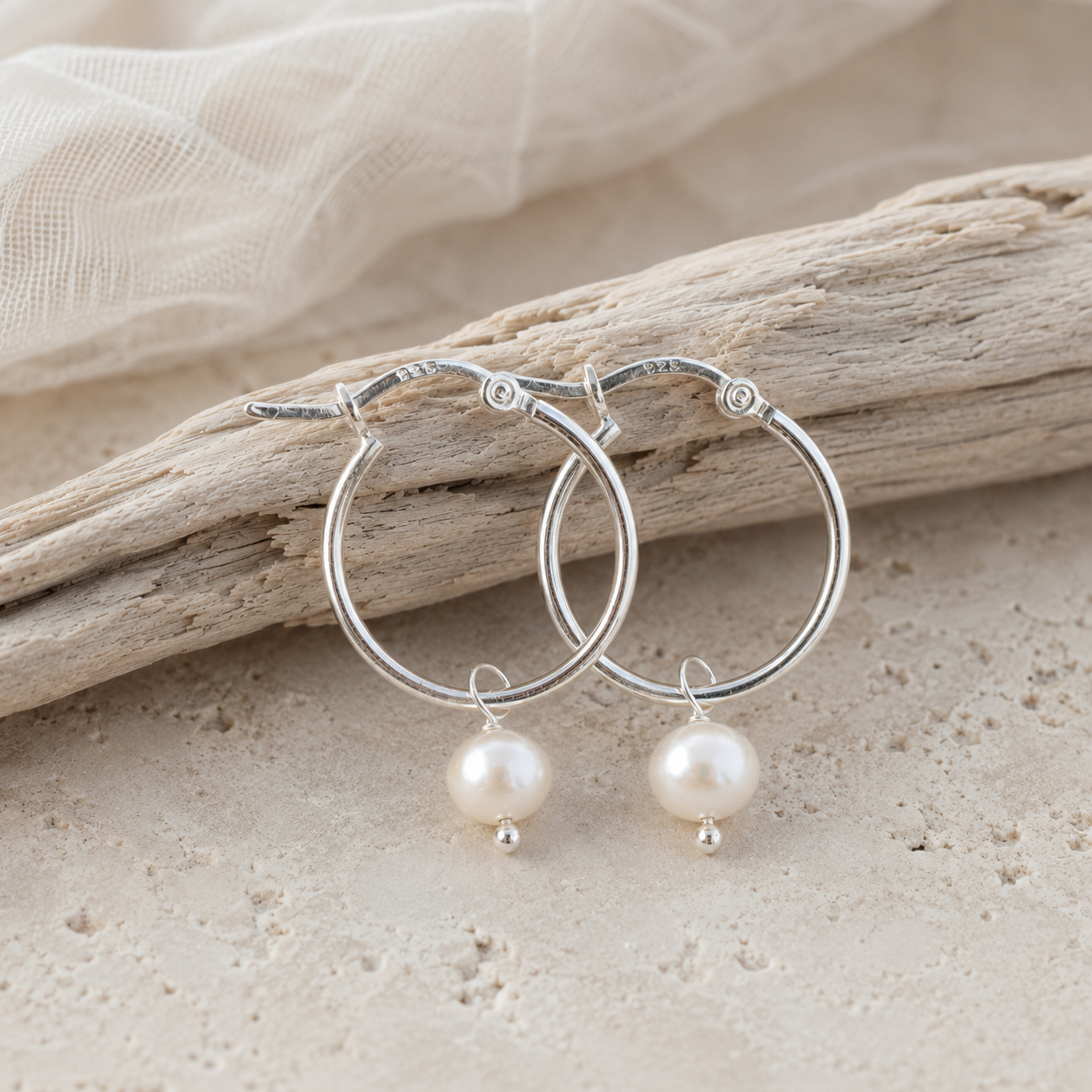 A pair of silver hoop earrings with dangling white pearl beads, displayed on a piece of driftwood over a light sandy surface.