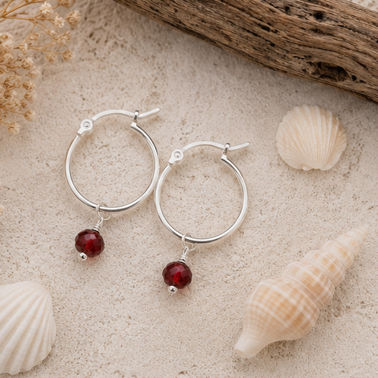 A pair of sterling silver hoop earrings with garnet gemstones, displayed on a sandy surface surrounded by seashells and dried flowers.