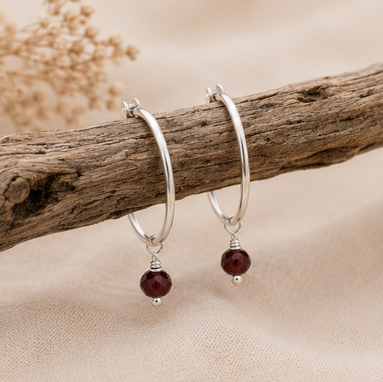 A pair of sterling silver hoop earrings with small dangling semi precious garnet faceted beads displayed on a rustic wooden branch over a soft beige background.