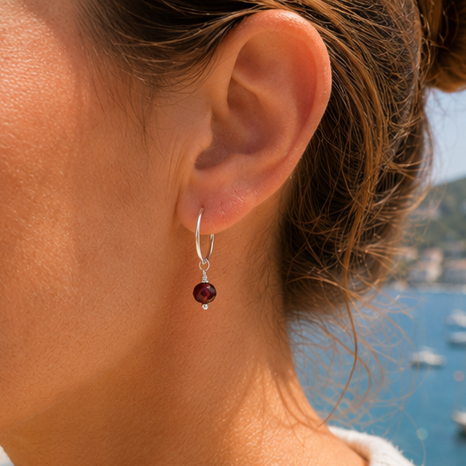 A close-up of a woman's ear wearing a silver hoop earring with a dangling garnet red gemstone, set against a blurred background of blue water and coastal scenery.