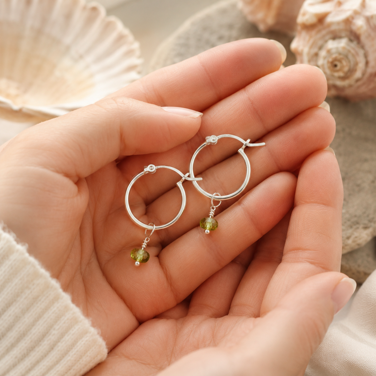 A pair of silver hoop earrings with peridot bead drops, held in a person's hand with seashells in the background.
