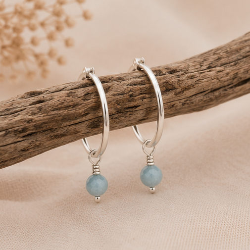 A pair of silver hoop earrings with aquamarine gemstones displayed on a piece of natural wood against a soft beige fabric background.