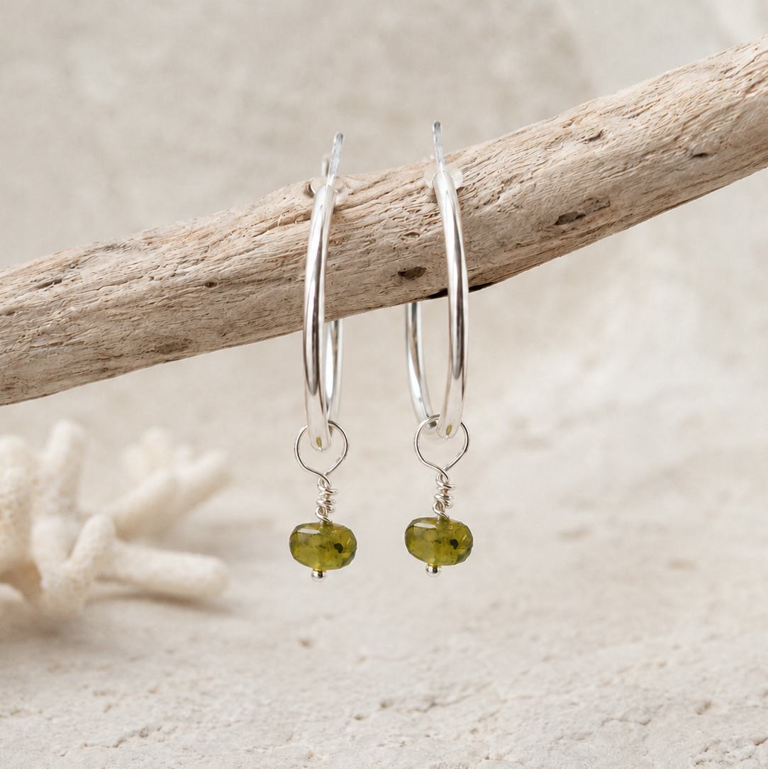 A pair of sterling silver hoop earrings with Peridot gemstone faceted bead drops, hanging from a piece of driftwood on a sandy beach background.